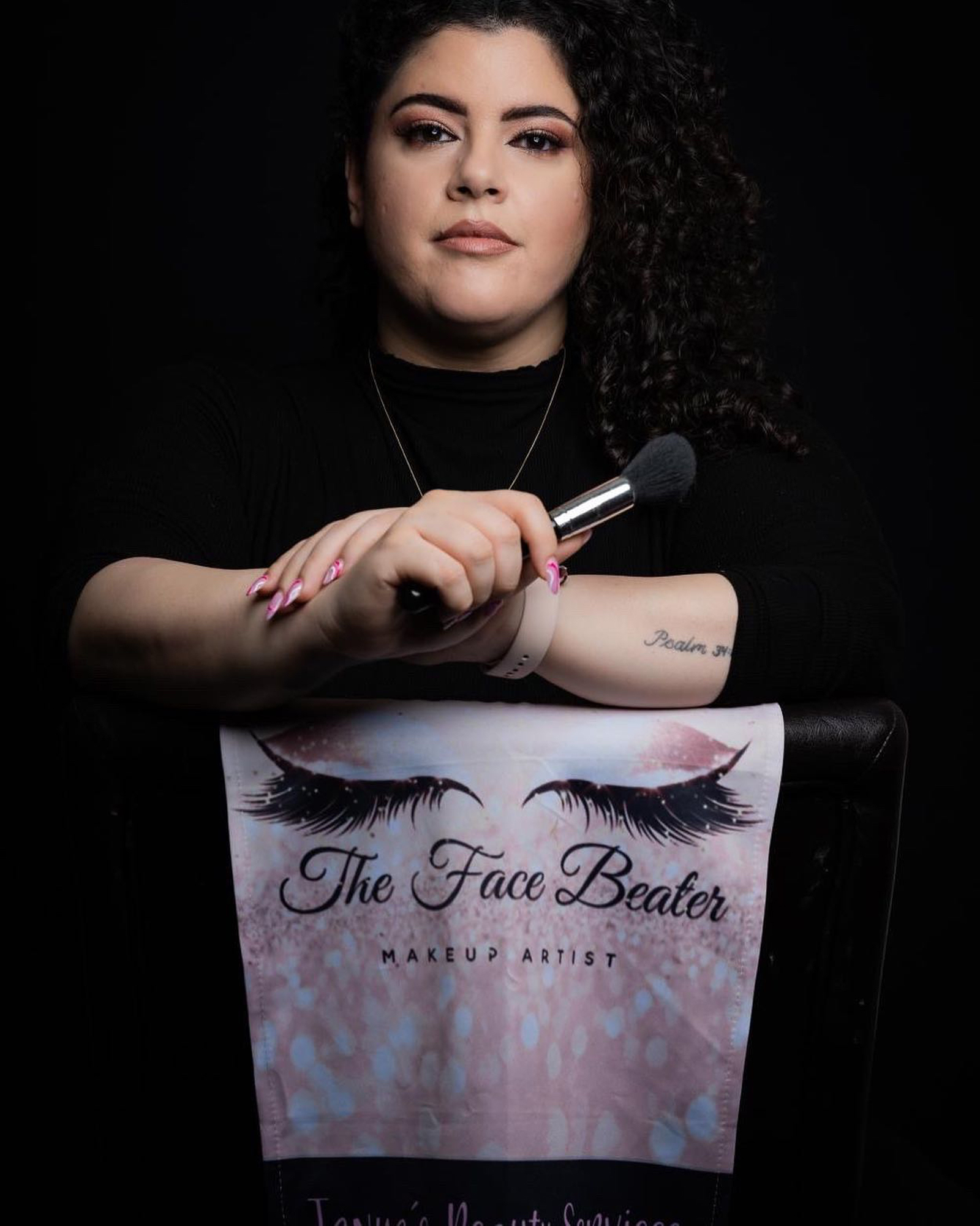 Tanya Garboushian holding a makeup brush, seated behind her branded chair cover reading The Face Beater, Makeup Artist