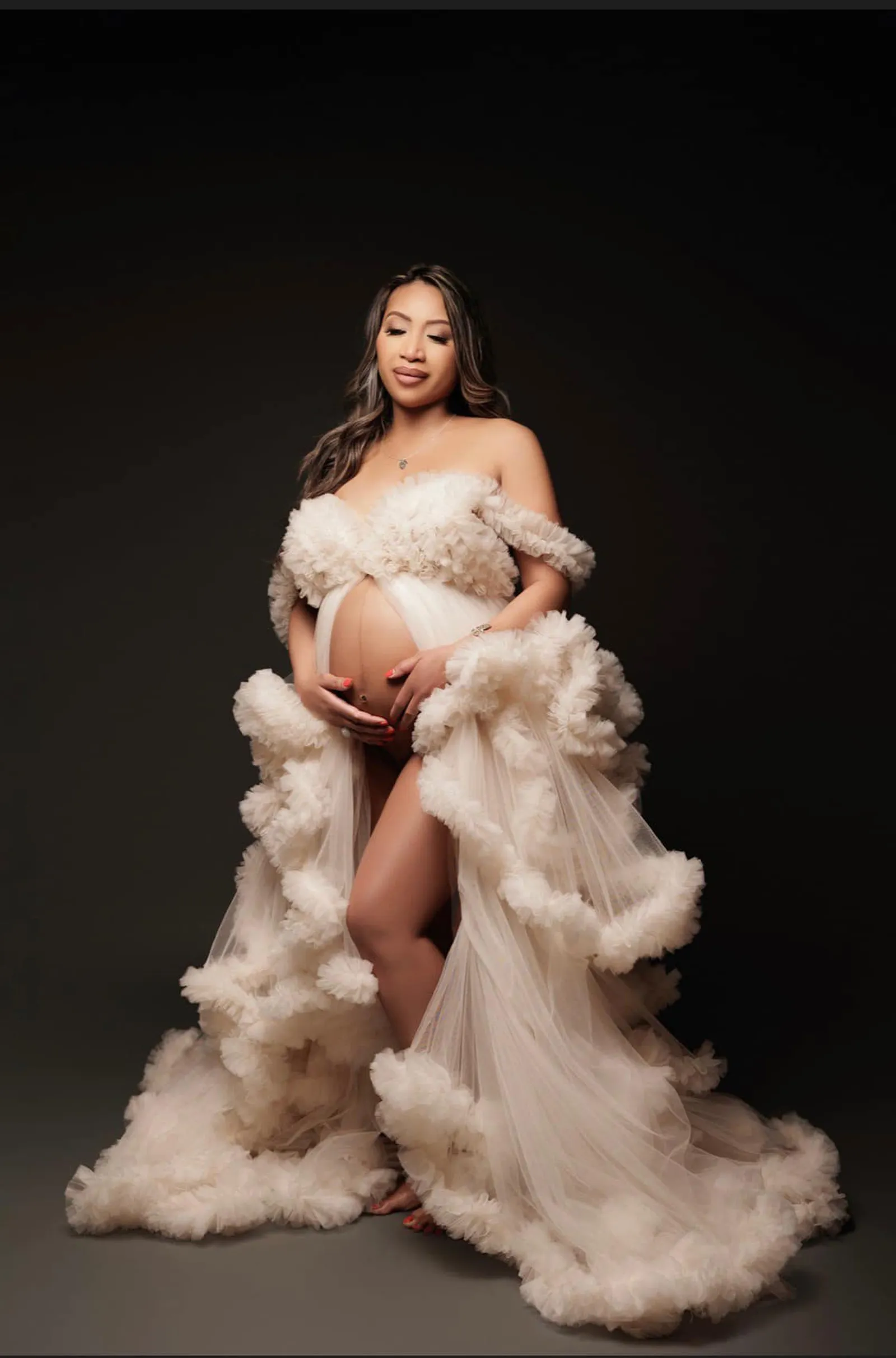 A maternity portrait: a client in a cream tulle gown with soft glowing makeup on a dark studio background