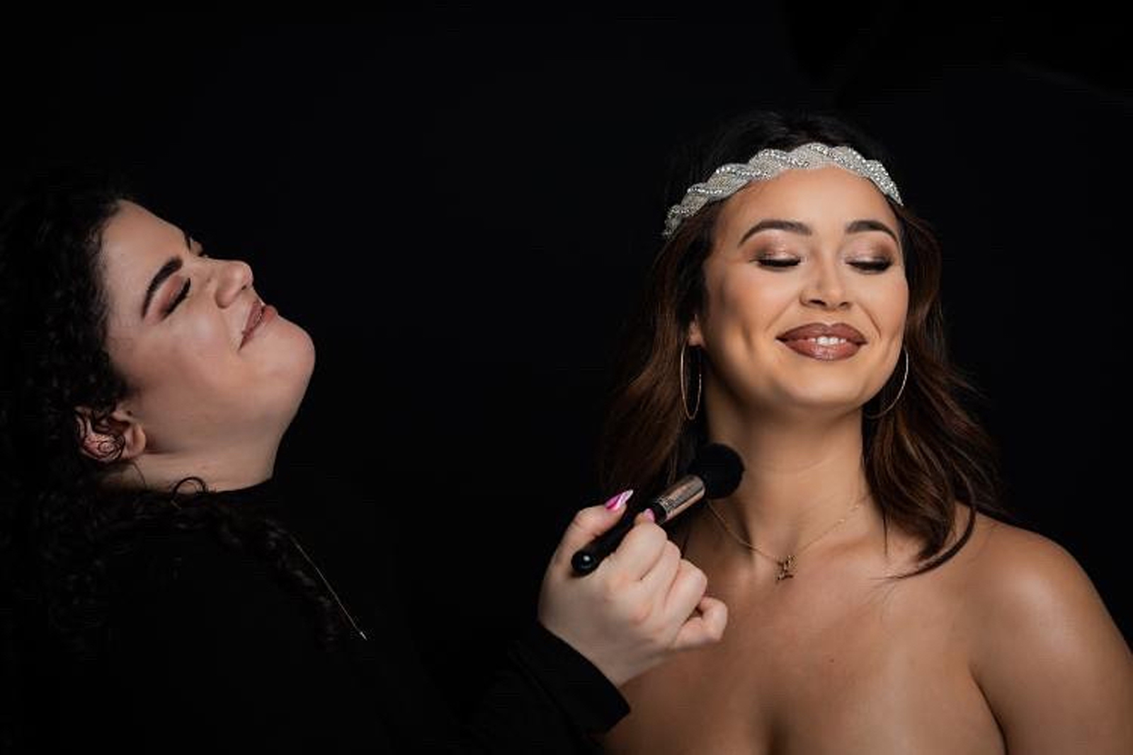Tanya laughing as she sweeps a brush across a bride's collarbone, both lit against black